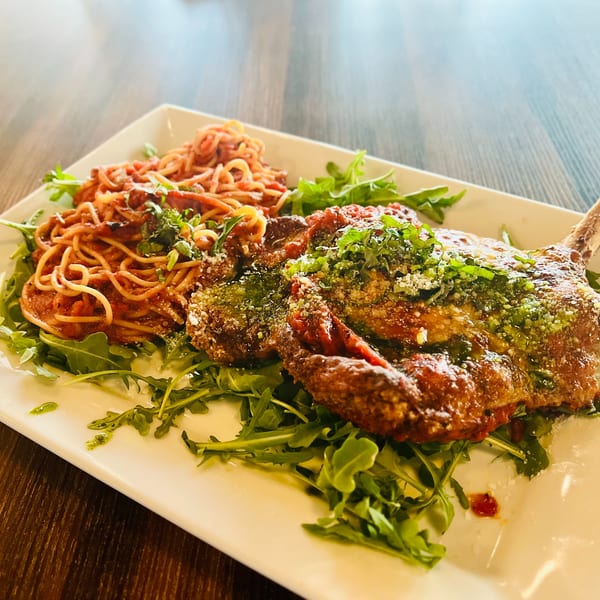Veal chop parmesan with spaghetti and arugula on a white rectangular plate.