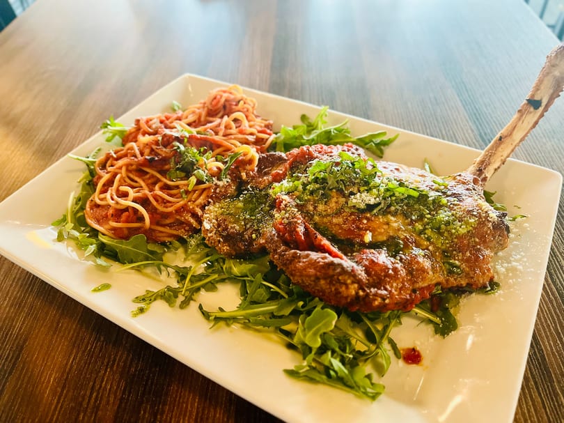 Veal chop parmesan with spaghetti and arugula on a white rectangular plate.