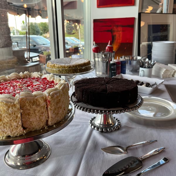 A dessert buffet with three cakes, plates, and serving utensils on a white tablecloth.