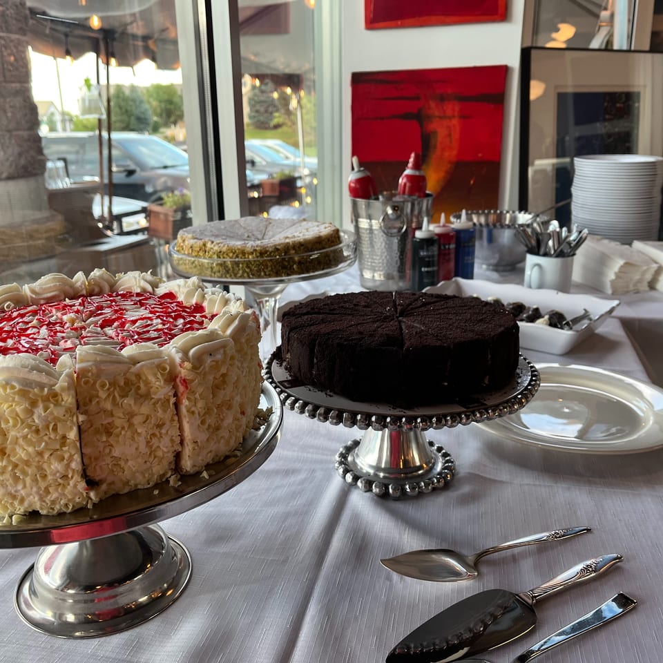A dessert buffet with three cakes, plates, and serving utensils on a white tablecloth.