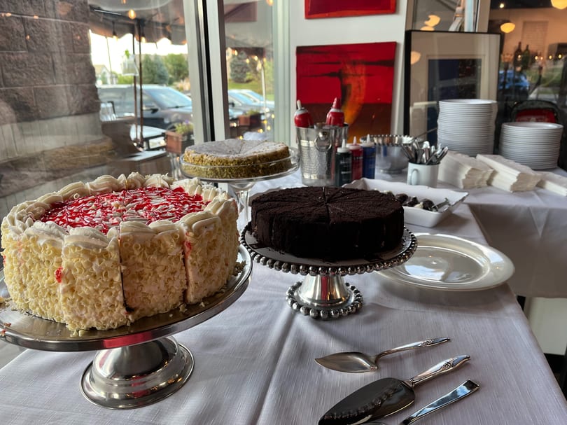 A dessert buffet with three cakes, plates, and serving utensils on a white tablecloth.