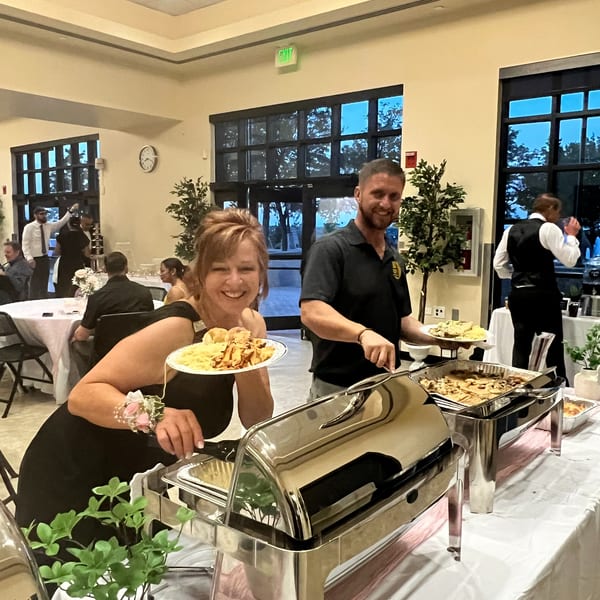 A woman smiles while holding a plate of food from a buffet, with a man serving behind her.