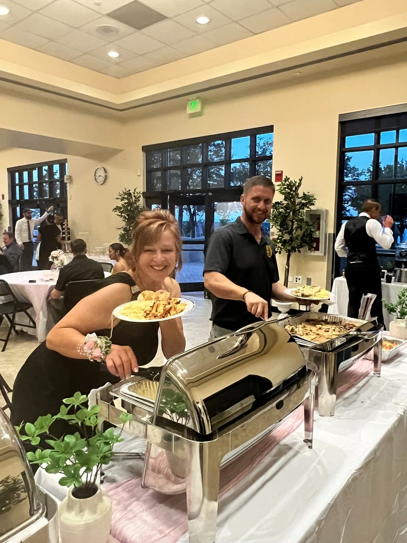 A woman smiles while holding a plate of food from a buffet, with a man serving behind her.