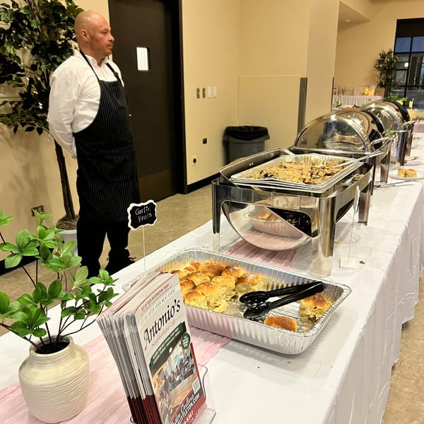 A chef stands beside a buffet table with chafing dishes, garlic knots, and Antonio's Cafe brochures.