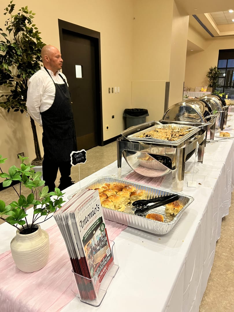 A chef stands beside a buffet table with chafing dishes, garlic knots, and Antonio's Cafe brochures.